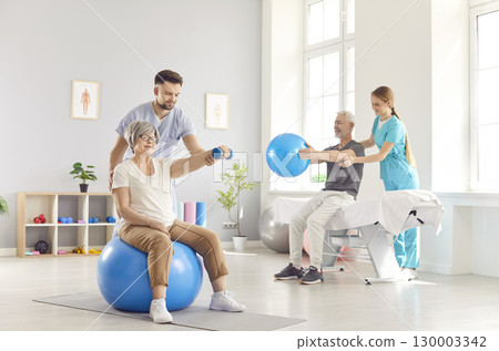 Health care workers helping mature people to do sport exercises with dumbbells and fit ball in gym. 130003342