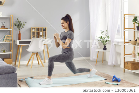 Active sporty woman doing front lunges on yoga mat at home, having fitness workout for body strength 130003361