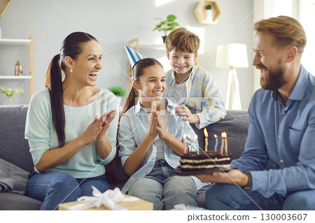 Happy girl in party hat celebrating birthday with family at home, looking at cake with candles Happy girl in party hat celebrating birthday with family at home, looking at cake with candles 130003607