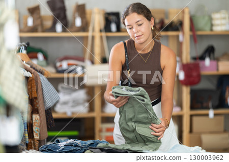 Girl customer choose trousers in store, and buyer carefully examines product. 130003962