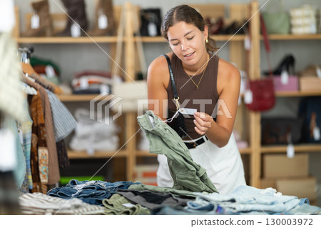 Girl customer choose trousers in store, and buyer carefully examines product. 130003972