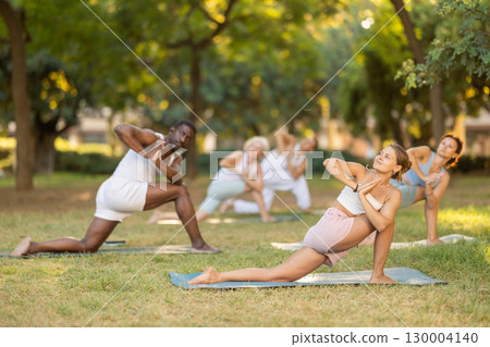 Multinational yoga enthusiasts with female coach perform Ardha Parshvottanasana in park 130004140