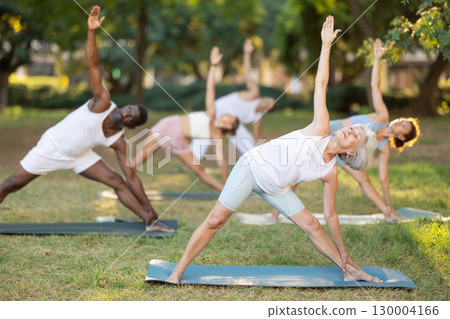Multinational yoga enthusiasts with mature female coach perform Trikonasana in park 130004166
