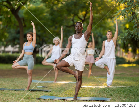African American man doing Vrikshasana during group yoga in park African American man doing Vrikshasana during group yoga in park 130004298