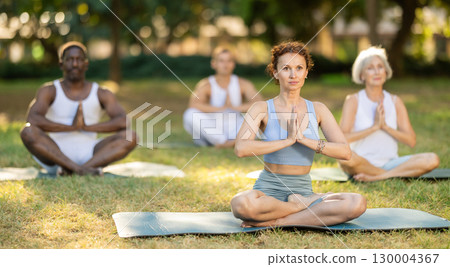 Multinational yoga enthusiasts with female coach perform padmasana in park 130004367