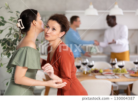 Smiling woman hostess hugging and kissing guest at dinner party 130004387