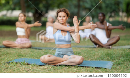 Portrait of women and men sitting on gym mat and doing yoga meditation in lotus position and arms raised in park Portrait of women and men sitting on gym mat and doing yoga meditation in lotus position and arms raised in park 130004392