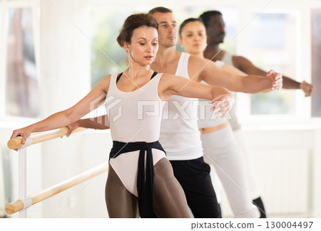 Ballerina performing battement tendu jete at ballet barre at group class 130004497