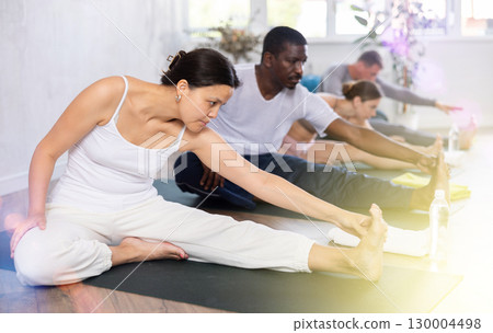 Portrait of woman making stretching legs with multinational group sitting on sports mat and making yoga exercise at fitness club 130004498
