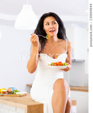 Woman sitting on kitchen table and eating salad Woman sitting on kitchen table and eating salad 130004644