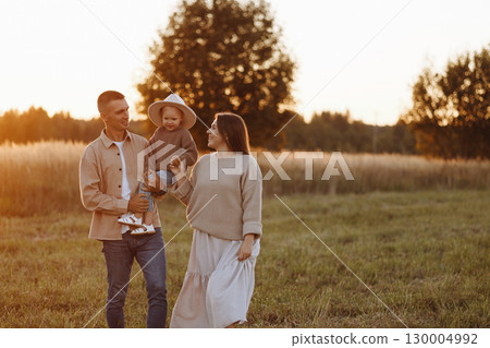 A family of three, a man and a woman with a child in their arms, stand in a field at sunset 130004992