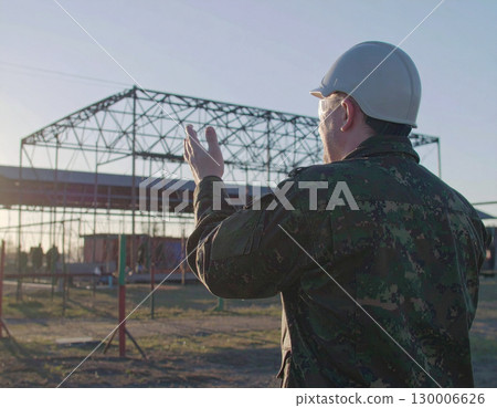 AI generated A uniformed soldier stands at the frame edge gesturing toward an active military training camp, showcasing discipline and teamwork. The background features obstacle courses and barracks 130006626