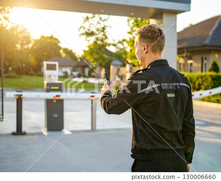 AI generated A professional security guard stands at the frame edge gesturing toward a guarded checkpoint with security booth barrier. The scene conveys safety, access control property protection. 130006660