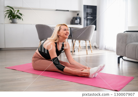Focused blonde middle aged woman balancing on one leg while stretching the other forward for flexibility and strength during home workout in modern living room 130007907