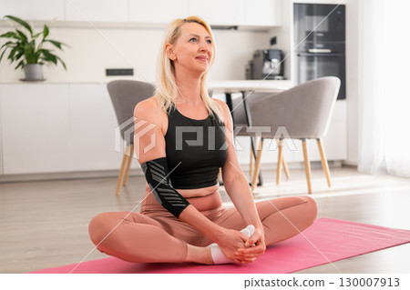 Calm middle aged blonde woman sitting in lotus pose practicing yoga meditation in stylish bright kitchen of modern apartment 130007913