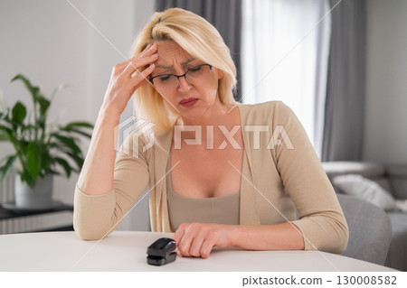 Woman in beige outfit sitting at home with modern interior, holding pulse monitor and appearing uncomfortable due to headache 130008582