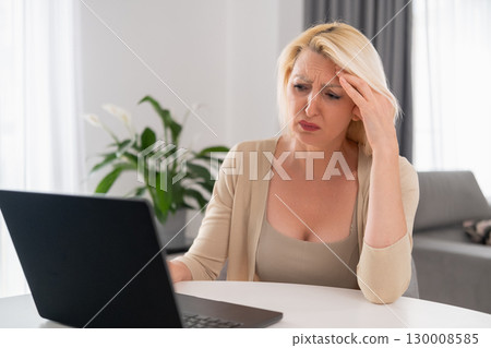 Middle aged blonde woman in beige clothes sadly looking at bill, sitting in cozy living room, concept of financial problems and household stress Middle aged blonde woman in beige clothes sadly looking at bill, sitting in cozy living room, concept of financial problems and household stress 130008585