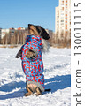 Small dog wearing a blue and red winter coat standing on hind legs in the snow 130011115