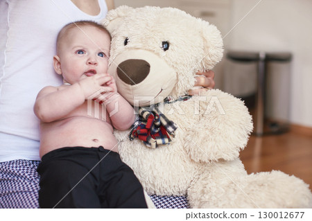 Four months old baby boy sitting on her mother knees, large plush teddy bear next to him 130012677