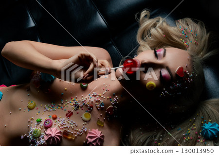 Woman with body art poses on a black surface surrounded by colorful sweets 130013950