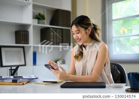 Smiling young businesswoman using smartphone at office desk with documents 130014574