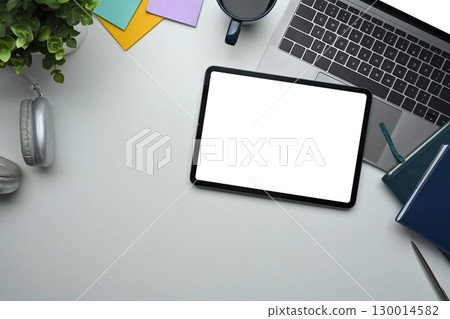 Top view of tablet with blank screen on desk alongside laptop, coffee cup, and notebooks 130014582