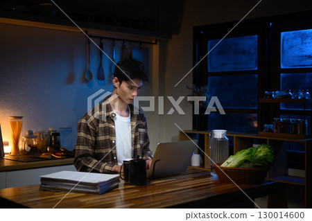 Concentrated freelancer sitting at kitchen counter, using laptop at midnight surrounded by documents 130014600