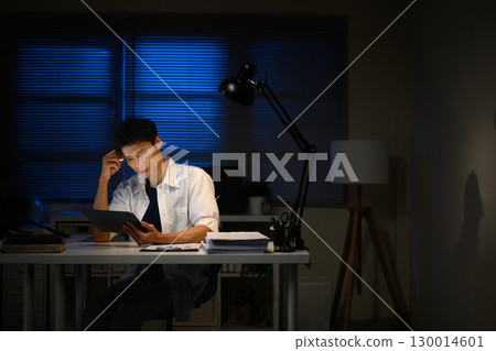 Focused man using digital tablet while working overtime under desk lamp Focused man using digital tablet while working overtime under desk lamp 130014601