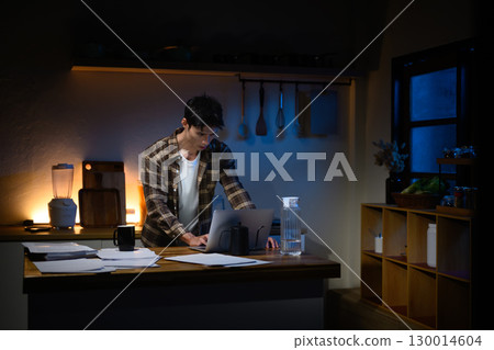 Young man working late night on laptop in a dimly lit kitchen, focusing on tasks with papers 130014604