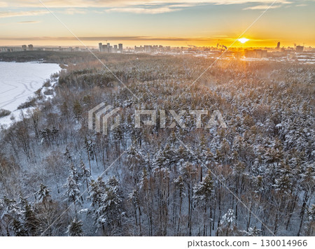 Snow-covered forest on lake shore with the first ice at sunset and the city on horizon, aerial view Snow-covered forest on lake shore with the first ice at sunset and the city on horizon, aerial view 130014966
