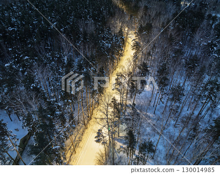 Aerial view of illuminated road in night winter forest with high pine or spruce trees covered by snow. Aerial view of illuminated road in night winter forest with high pine or spruce trees covered by snow. 130014985