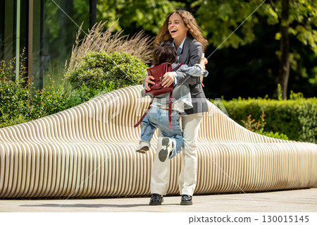 Joyful woman hugs child with backpack near stylish bench outdoor Joyful woman hugs child with backpack near stylish bench outdoor 130015145