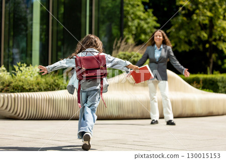 Excited schoolgirl running toward smiling woman with open arms holding notebook 130015153