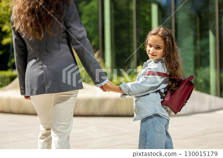 Woman with long hair walking with schoolgirl outdoors by glass facade 130015179