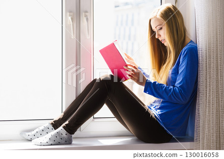 Woman sitting on window sill reading book at home 130016058