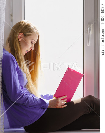 Woman sitting on window sill reading book at home Woman sitting on window sill reading book at home 130016059