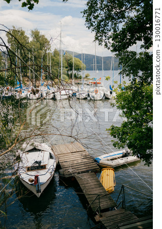 Sailboats are moored at wooden pier on calm lake, with lush green trees framing serene scene and rolling hills visible in distance under cloudy sky. Wooden pier hosting moored sailboats, cloudy sky  130016771
