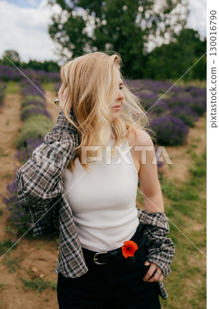 Blonde woman wearing casual clothes with poppy flower attached to her belt is posing in lavender field, touching her hair and looking away on sunny summer day. Blonde female standing in lavender field Blonde woman wearing casual clothes with poppy flower attached to her belt is posing in lavender field, touching her hair and looking away on sunny summer day. Blonde female standing in lavender field 130016790