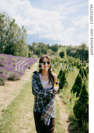 Beautiful young woman wearing sunglasses and casual clothes is walking near lavender flowers and holding smartphone on sunny summer day, enjoying beautiful nature and relaxing atmosphere on sunny day 130016794
