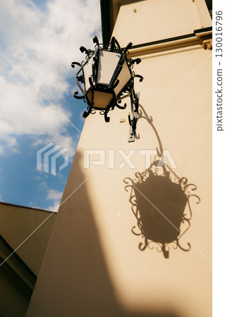 Ornate vintage street lamp attached to building casts distinct shadow on facade under vibrant blue sky, creating captivating interplay of light and design. Vintage street lamp casting shadow on wall Ornate vintage street lamp attached to building casts distinct shadow on facade under vibrant blue sky, creating captivating interplay of light and design. Vintage street lamp casting shadow on wall 130016796