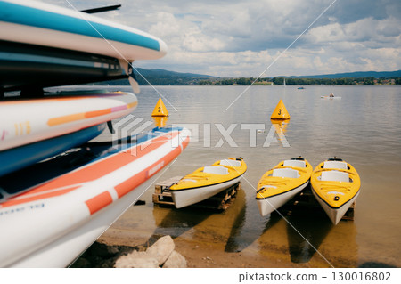 Yellow kayaks and colorful paddle boards are stored on shore of lake on partly cloudy summer day, while canoeist practices in distance. Colorful watercraft resting on lakeside shore next to sap boards 130016802
