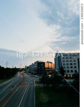 Empty road stretching towards modern office buildings and hotel, set against cloudy sunset sky in tranquil urban business district, creating serene and atmospheric scene. Vacant roadway in hushed zone 130016813