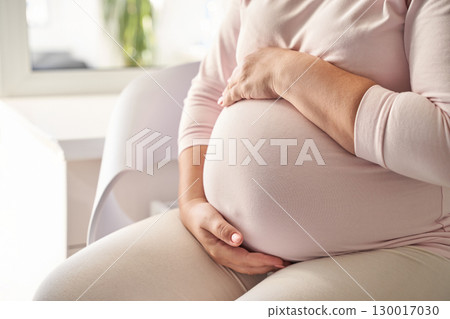 Young caucasian woman cradling pregnant belly in light pink outfit sitting 130017030