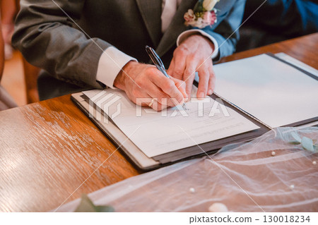 Hands of a person in formal attire signing a wedding document. The image captures a meaningful moment symbolizing commitment and legal union, set against a wooden surface Hands of a person in formal attire signing a wedding document. The image captures a meaningful moment symbolizing commitment and legal union, set against a wooden surface 130018234