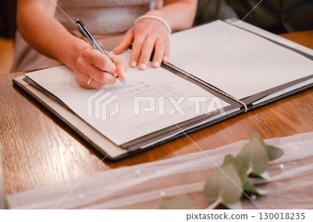 Person signing a document at a wooden table, expressing commitment and responsibility Firms grip on pen suggests focused action Document open on table with blank pages visible 130018235