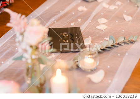 Two wedding rings placed on a decorative book surrounded by delicate flowers, candles, and petals on a beautifully arranged table, creating a romantic atmosphere for a special occasion 130018239