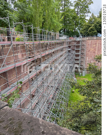 Extensive scaffolding installed along an ancient brick wall surrounded by lush greenery in a park. The scene illustrates the preparation for restoration or maintenance work 130019718