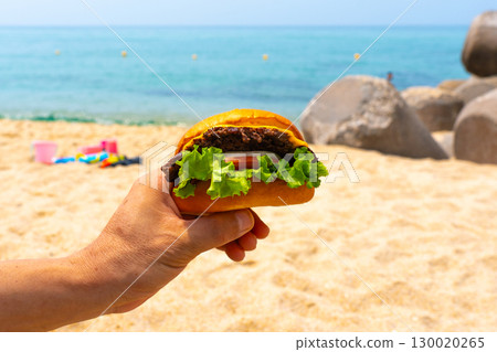 Man holding burger in a hand against beach background Man holding burger in a hand against beach background 130020265