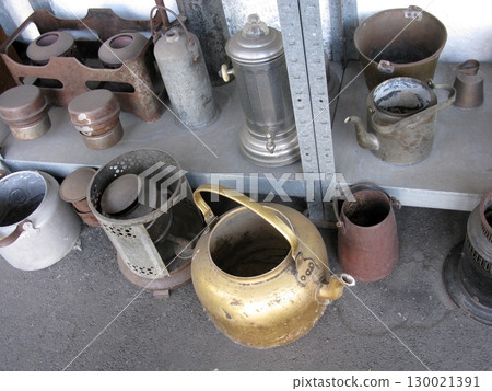 A cluttered shelf holding an array of vintage kitchenware, including a brass kettle and tin pots, showcasing rustic culinary tools from a bygone era 130021391