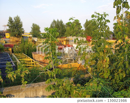 Overgrown Industrial Yard with Rusty Machinery and Lush Greenery in the Evening 130021986